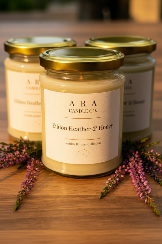Three glass jar candles with gold lids and cream labels are arranged on a wooden surface, with the front candle labeled 'ARA Candle Co. Eildon Heather & Honey' and purple heather sprigs around it.