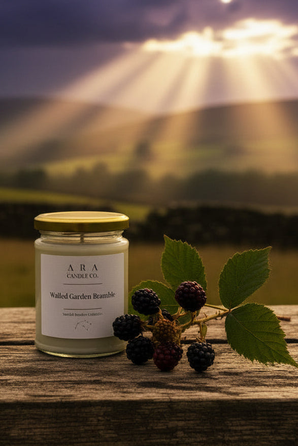 A glass jar candle with a gold lid and white label sits on a weathered wooden surface outdoors, styled with fresh blackberries and green leaves. The background shows a soft-focus rural landscape with rolling green hills under a cloudy sky.