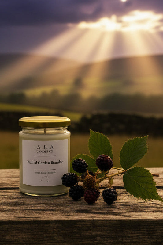 A glass jar candle with a gold lid and white label sits on a weathered wooden surface outdoors, styled with fresh blackberries and green leaves. The background shows a soft-focus rural landscape with rolling green hills under a cloudy sky.