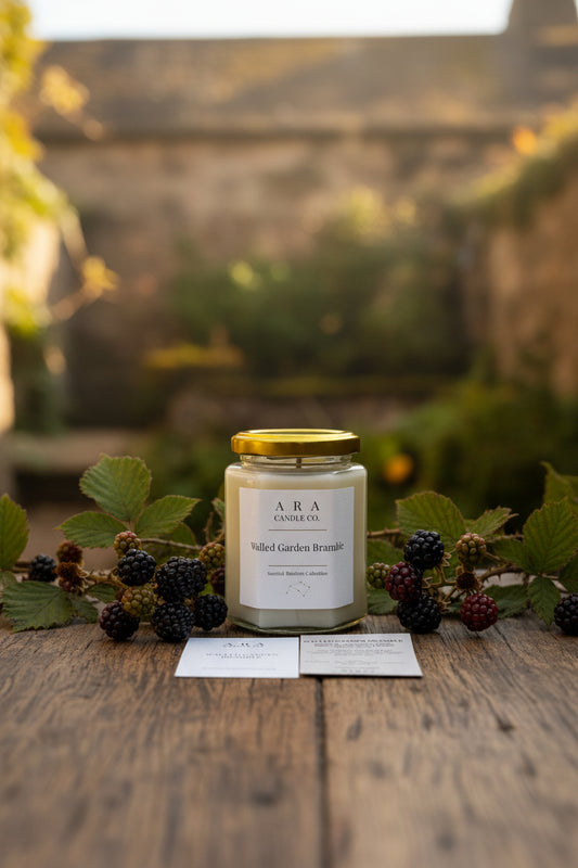 A glass jar candle with a gold lid and a white label reading 'ARA CANDLE CO. Walled Garden Bramble' sits on a wooden table, with two printed cards placed in front.