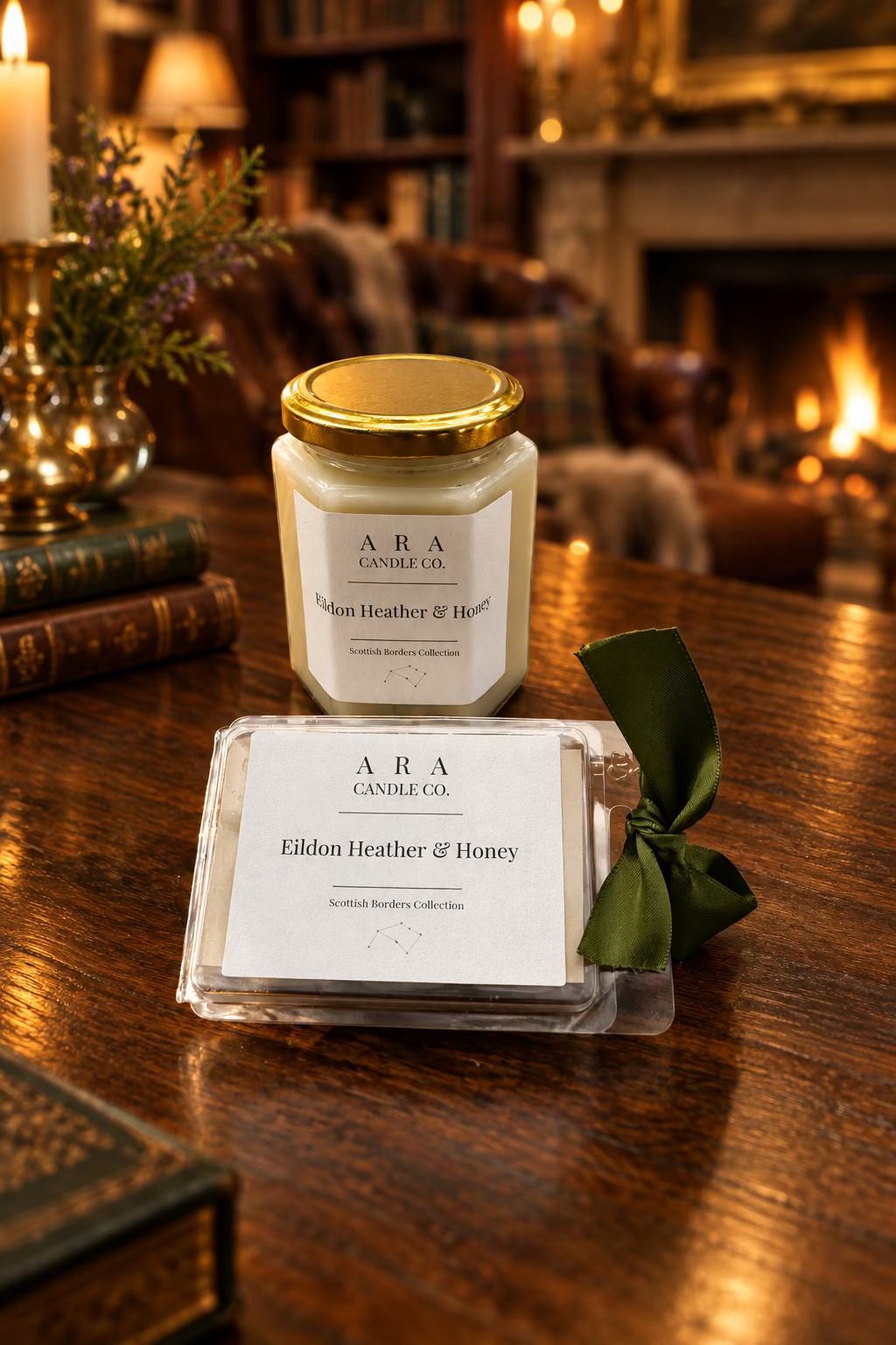 A jar candle and a packaged candle box from ARA Candle Co on a wooden table. The jar candle has a gold lid and cream-colored wax, while the box in front has a white label reading 'Eildon Heather & Honey' and is tied with a dark green ribbon.
